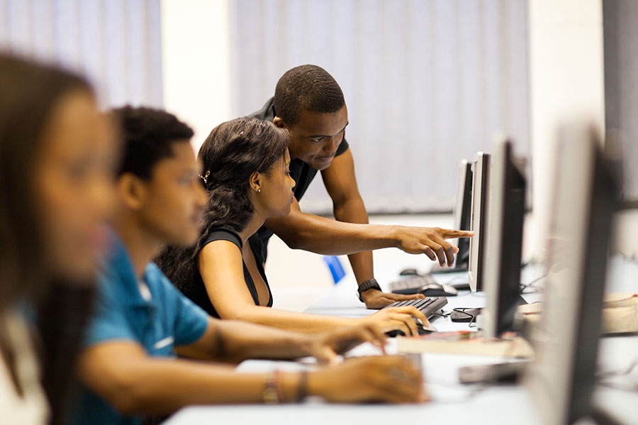 A coach pointing out something on screen while a participant works at a computer, with classmates at workstations nearby